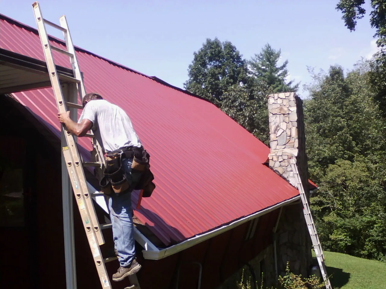 Roofer on a ladder beside a red metal roof and stone chimney — Soffit & Fascia Repair by Roofing Brothers in Brandywine